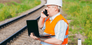 Network Rail job cuts could have “devastating consequences” for rail safety, warns TSSA Rail worker in orange high vis trackside with laptop. From Canva