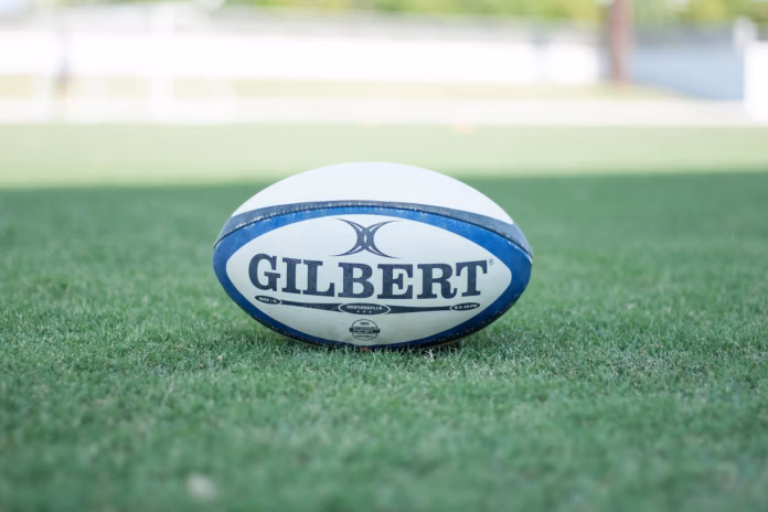 Rugby Ball A photo of a Rugby ball with the text "GILBERT" on it underlined. It is in the middle of a rugby pitch.